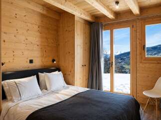 Experience cozy mountain living in this charming wooden bedroom, featuring a comfortable bed and stunning panoramic views of snow-capped peaks through large windows.
