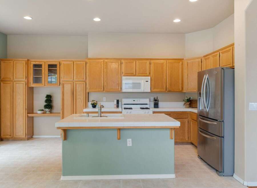 This spacious kitchen features light wood cabinetry, a large island with a tiled countertop, and a stainless steel refrigerator.