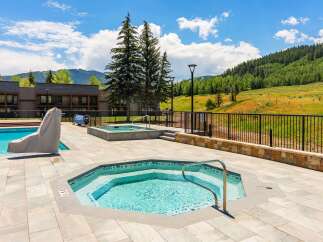 Enjoy a refreshing dip in the sparkling blue pool or relax in the inviting hot tub, both set against a backdrop of lush green mountains and a clear blue sky.