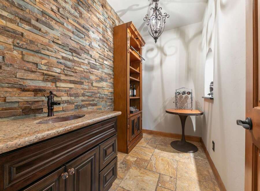 This stylish wet bar area features a striking stacked stone accent wall, complemented by a granite countertop and a dark wood vanity with ornate hardware.