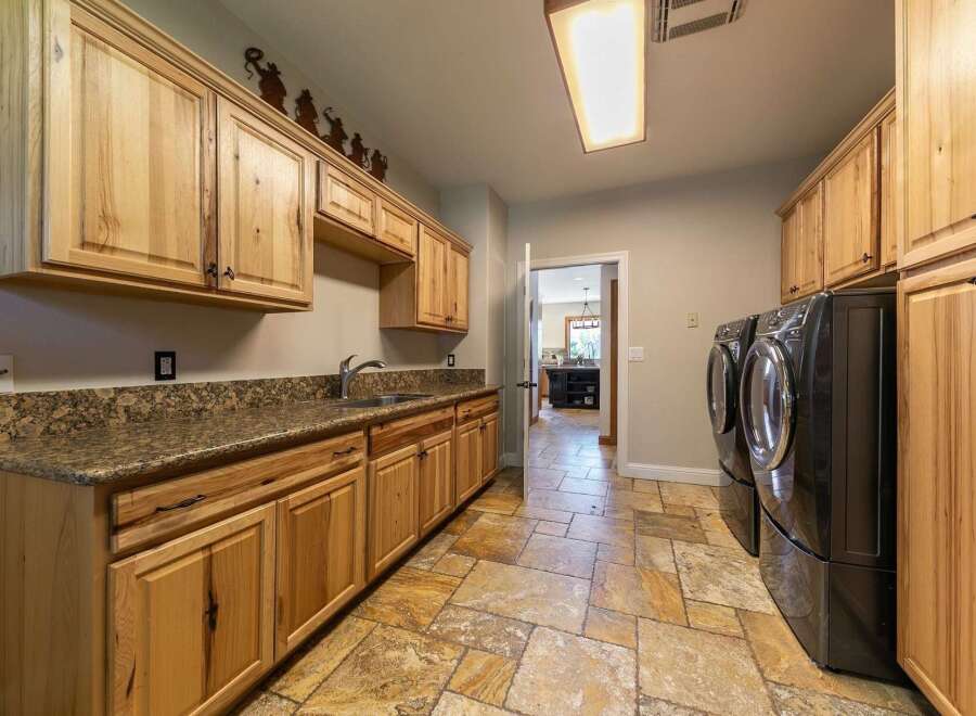 This spacious laundry room features ample natural wood cabinetry providing abundant storage, complemented by a granite countertop and a deep sink.