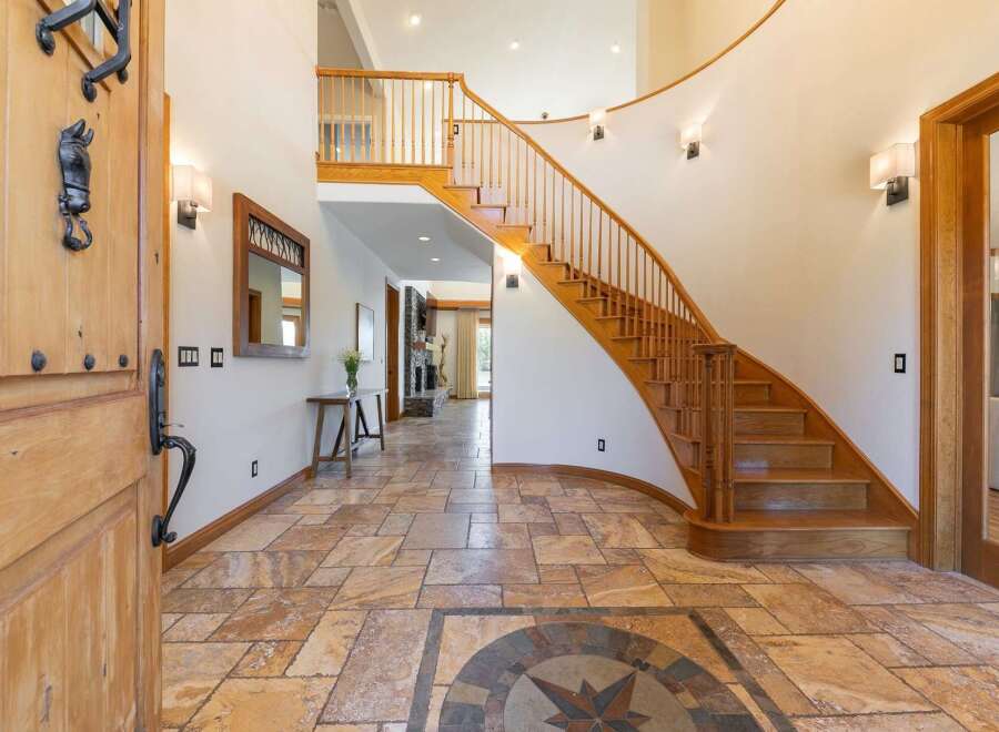 Step into this grand foyer featuring a stunning curved staircase with rich wooden railings and a decorative compass rose inlay on the travertine tile floor.