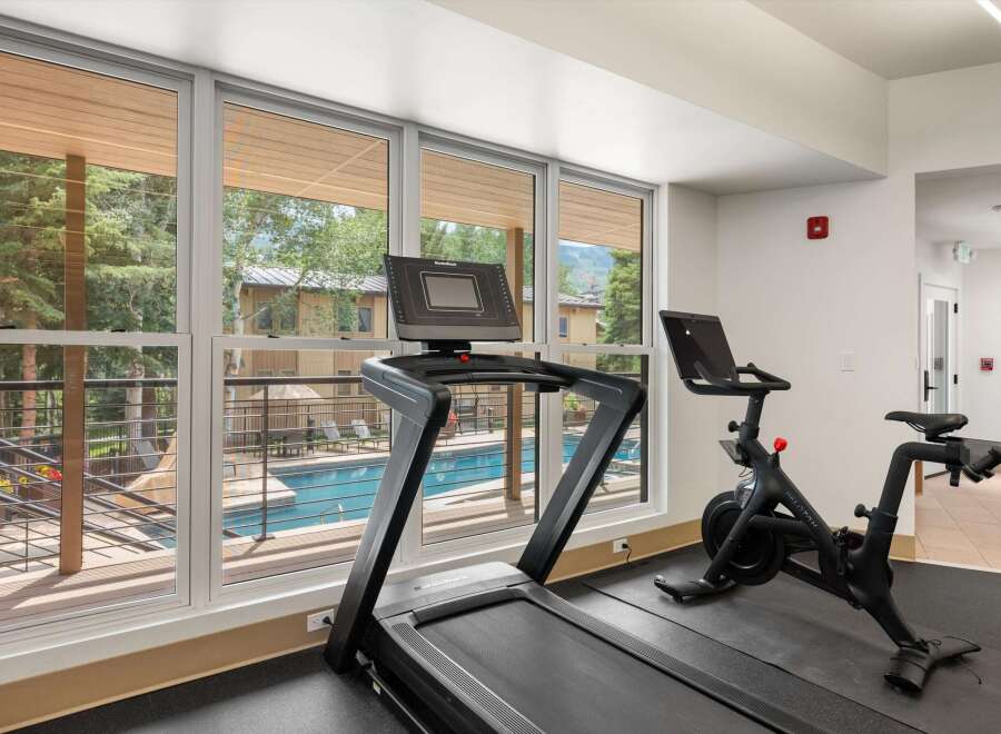 Elevate your fitness routine in this modern gym, featuring a treadmill and a Peloton bike with stunning views of the outdoor pool and surrounding trees.