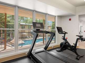 Elevate your fitness routine in this modern gym, featuring a treadmill and a Peloton bike with stunning views of the outdoor pool and surrounding trees.