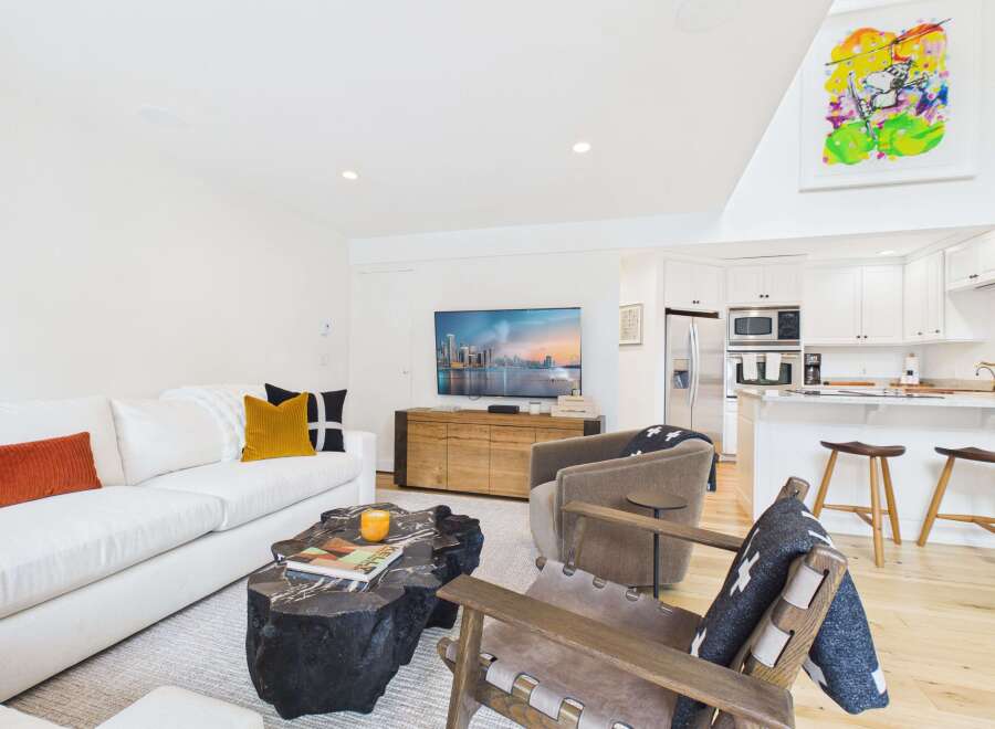This modern living room and kitchen area features a comfortable white sectional sofa adorned with accent pillows, a unique live-edge coffee table, and a large flat-screen TV.
