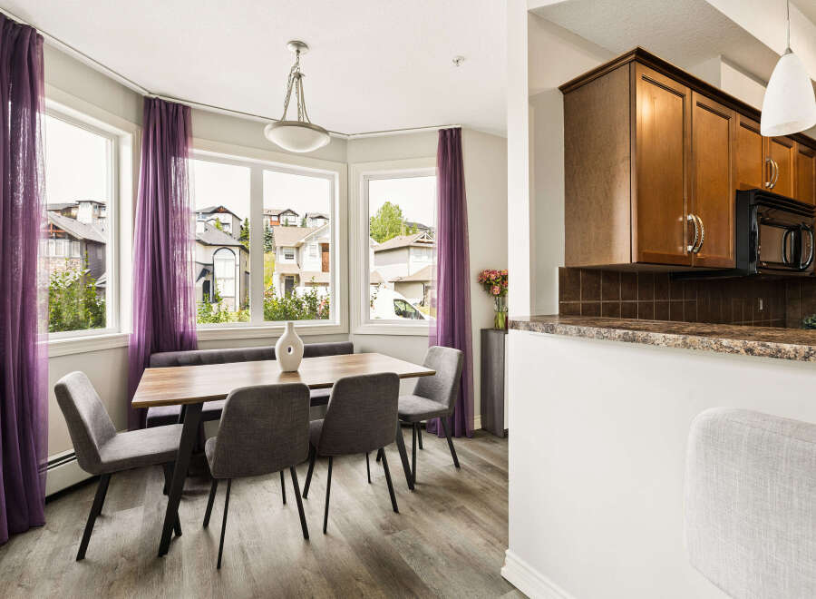 Bright and airy dining area featuring a modern wooden table with upholstered chairs and a bench, complemented by elegant purple sheer curtains framing large windows.