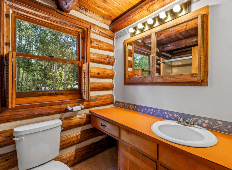 Experience rustic charm in this log cabin bathroom, featuring a bright orange vanity and a large window overlooking a serene forest.