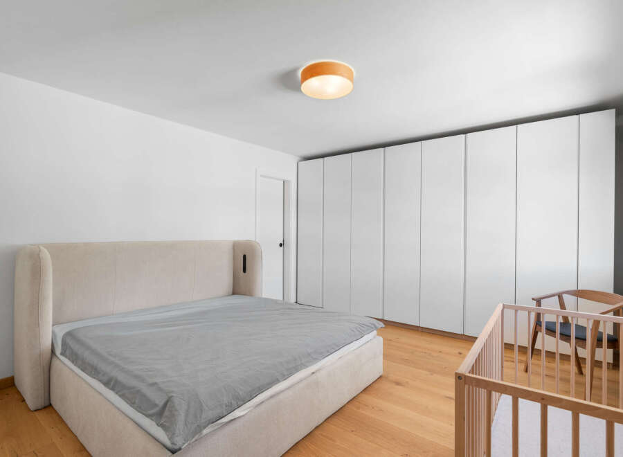 This modern bedroom features a spacious king-size bed with a plush upholstered headboard, complemented by a minimalist crib and a wooden accent chair.