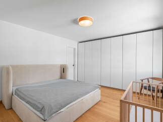 This modern bedroom features a spacious king-size bed with a plush upholstered headboard, complemented by a minimalist crib and a wooden accent chair.