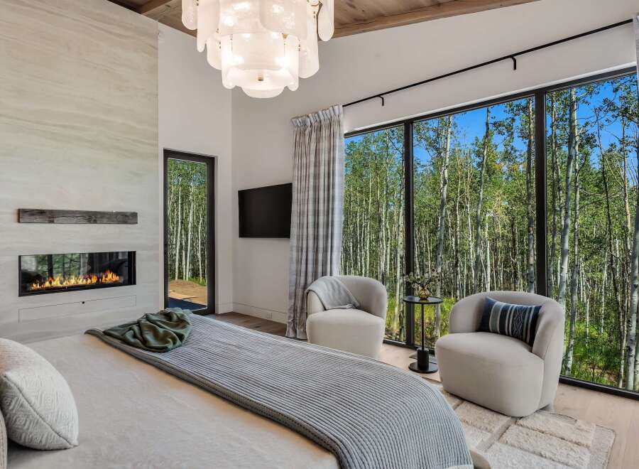 Experience luxury living with this stunning bedroom featuring a modern fireplace and expansive windows that offer breathtaking views of a serene forest.