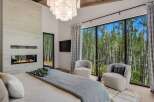 Experience luxury living with this stunning bedroom featuring a modern fireplace and expansive windows that offer breathtaking views of a serene forest.