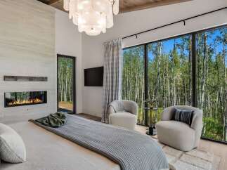 Experience luxury living with this stunning bedroom featuring a modern fireplace and expansive windows that offer breathtaking views of a serene forest.