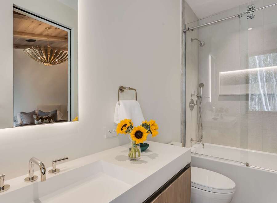 Experience modern luxury in this stunning bathroom featuring a sleek white vanity with contemporary chrome fixtures and a large, illuminated mirror reflecting a stylish bedroom.