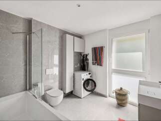 This modern bathroom features a sleek bathtub with a glass shower screen, a wall-mounted toilet, and elegant grey marble-effect tiles.