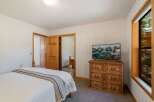 This charming bedroom features a comfortable bed with a striped throw, a wooden dresser topped with a flat-screen TV displaying a scenic landscape, and a large window offering views of the surrounding natural beauty.