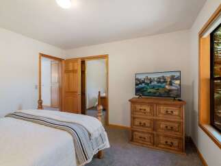 This charming bedroom features a comfortable bed with a striped throw, a wooden dresser topped with a flat-screen TV displaying a scenic landscape, and a large window offering views of the surrounding natural beauty.