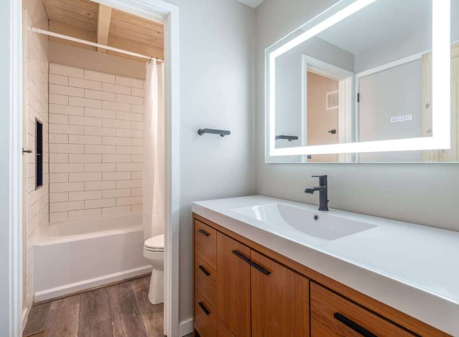 This modern bathroom features a sleek vanity with a white countertop and a black faucet, complemented by a large, illuminated mirror.