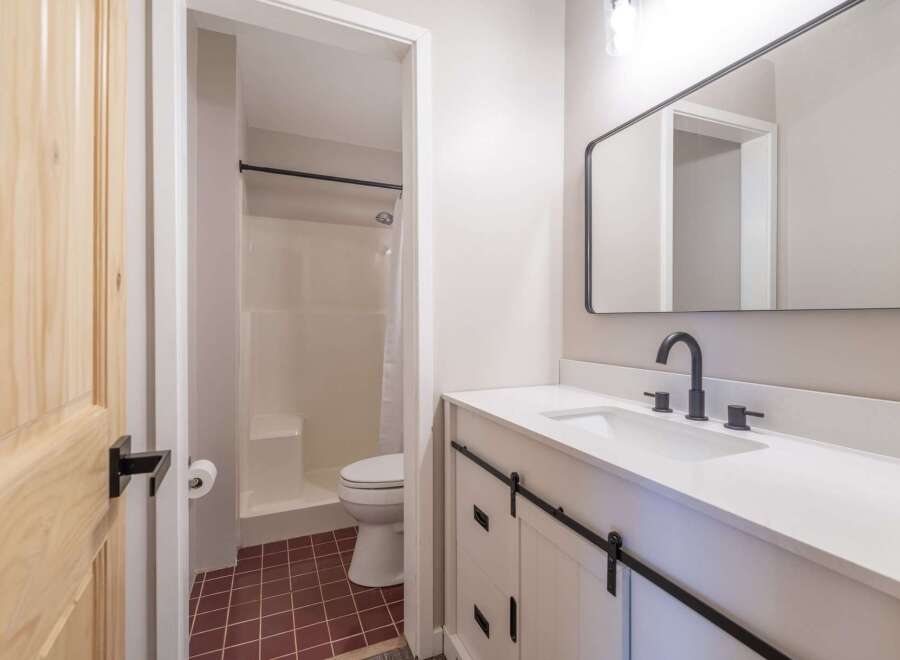 This modern bathroom features a sleek white vanity with barn-style doors and a black faucet, complemented by a large rectangular mirror.