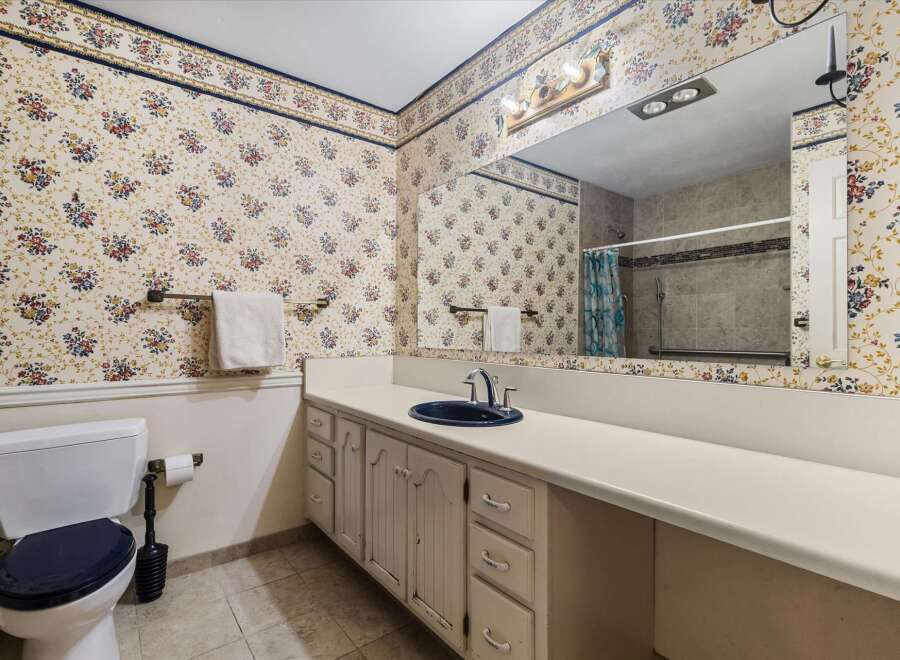 This charming bathroom features a floral wallpaper with a blue and yellow border, complemented by a white vanity with a navy sink and chrome fixtures.