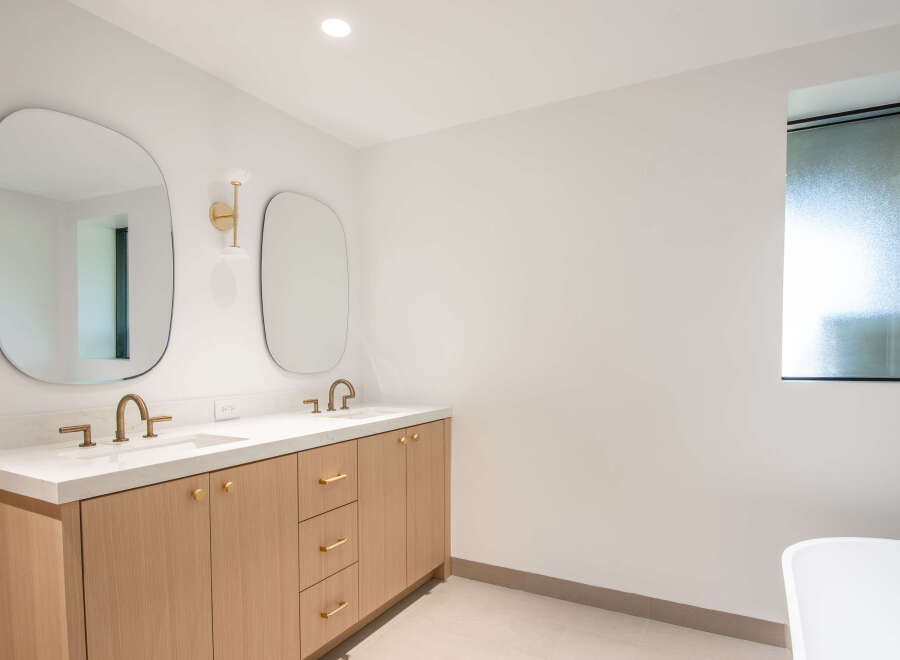 This modern bathroom features a double vanity with sleek gold fixtures and a light wood cabinet.