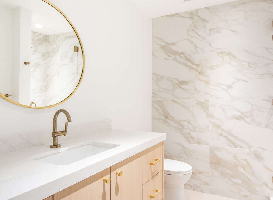 This modern bathroom features a sleek vanity with a white countertop and light wood cabinetry, complemented by a round gold-framed mirror.