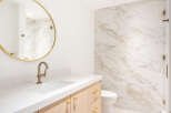 This modern bathroom features a sleek vanity with a white countertop and light wood cabinetry, complemented by a round gold-framed mirror.