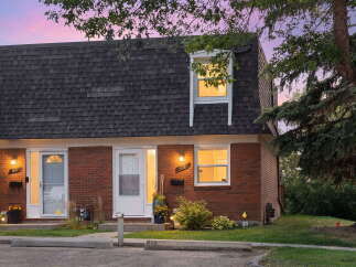 Charming brick townhouses with dark shingled roofs are illuminated by warm exterior lights under a vibrant twilight sky.