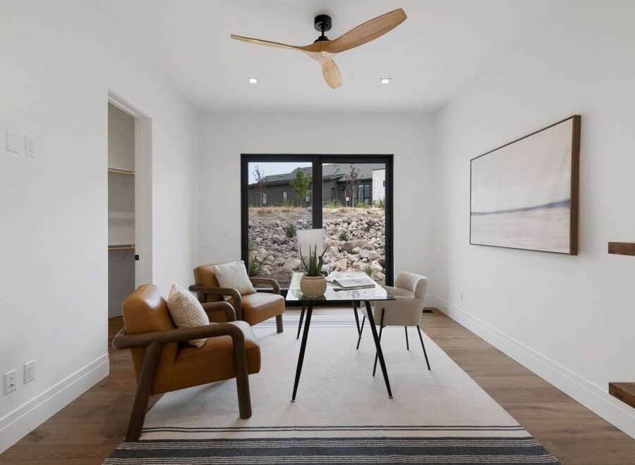 This modern office space features a sleek glass-top desk with black metal legs, complemented by two stylish tan leather armchairs with wooden accents.