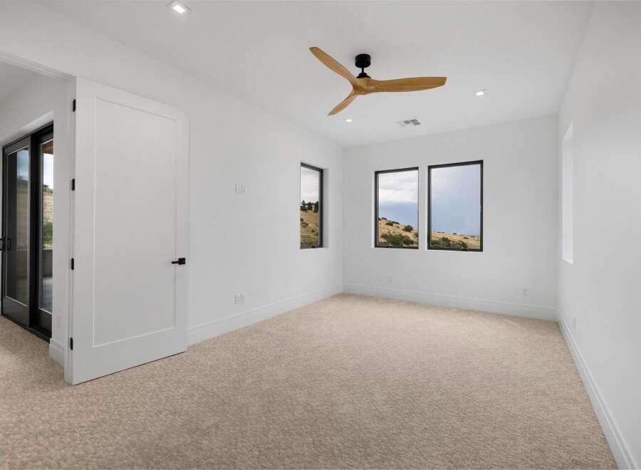 Experience modern living in this spacious bedroom featuring a sleek ceiling fan and large windows that offer breathtaking views of the surrounding landscape.