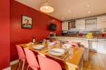 This inviting dining area features a warm red accent wall, a large wooden dining table set for a meal, and a modern kitchen with white cabinetry and stainless steel appliances.