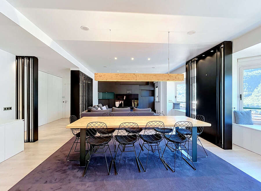 Experience modern living in this spacious open-concept dining and living area, featuring a striking wooden beam chandelier above a large dining table with contemporary wire chairs.