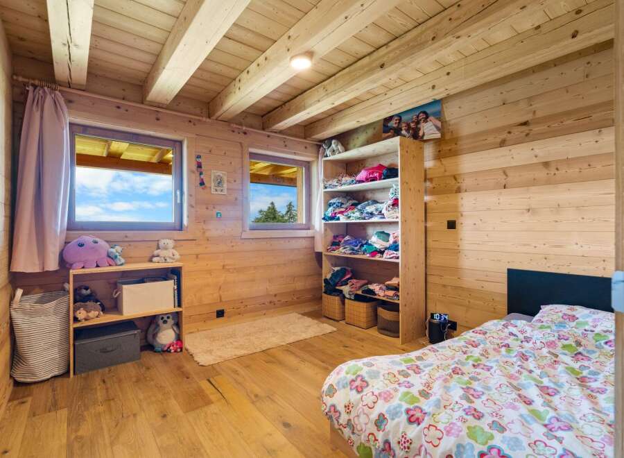 This charming wooden cabin bedroom features a cozy bed with a colorful duvet and ample storage solutions, including a tall shelving unit filled with clothes and a smaller bookshelf with toys.