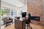 This home office features a rustic live-edge desk paired with a comfortable black leather office chair, set against a striking exposed brick wall with a modern fireplace.