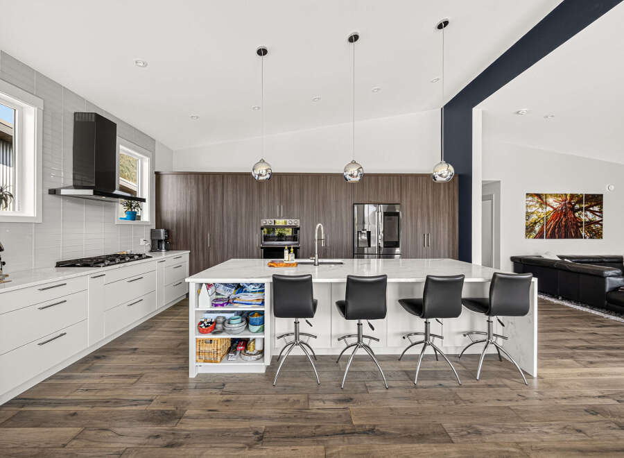 Experience modern luxury in this stunning kitchen, featuring sleek white cabinetry, a spacious island with waterfall countertops, and a sophisticated dark wood backsplash.