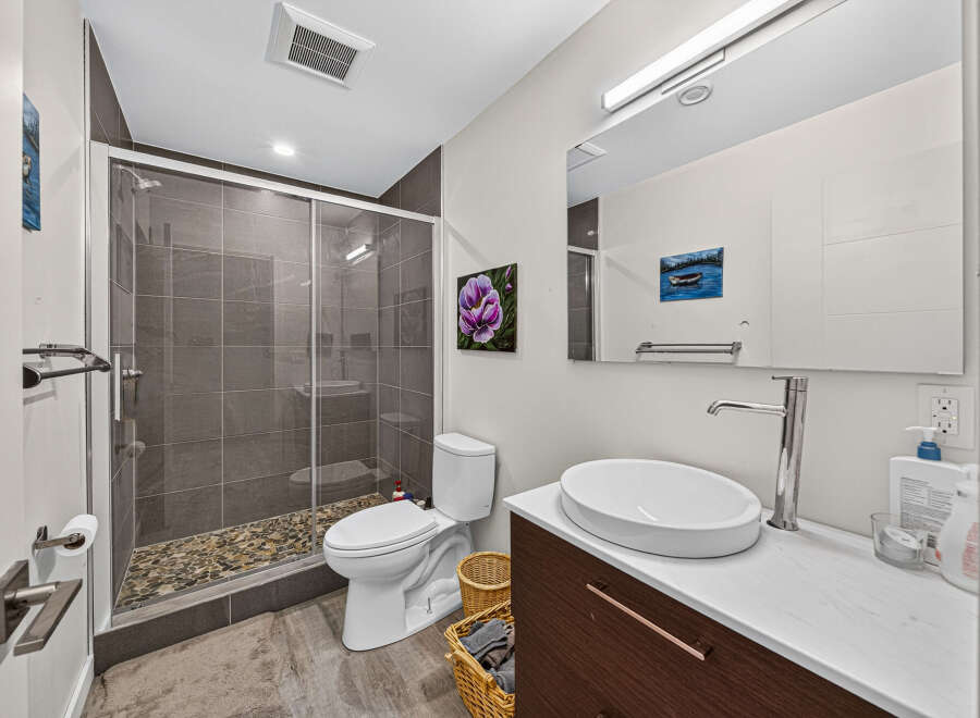 This modern bathroom features a sleek walk-in shower with pebble flooring and grey tiled walls, complemented by a contemporary white toilet and a stylish vessel sink vanity.