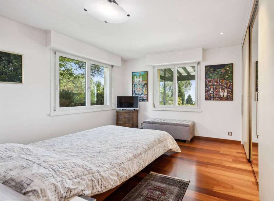 This charming bedroom features a comfortable bed with a patterned duvet, complemented by large windows offering picturesque views of lush greenery.