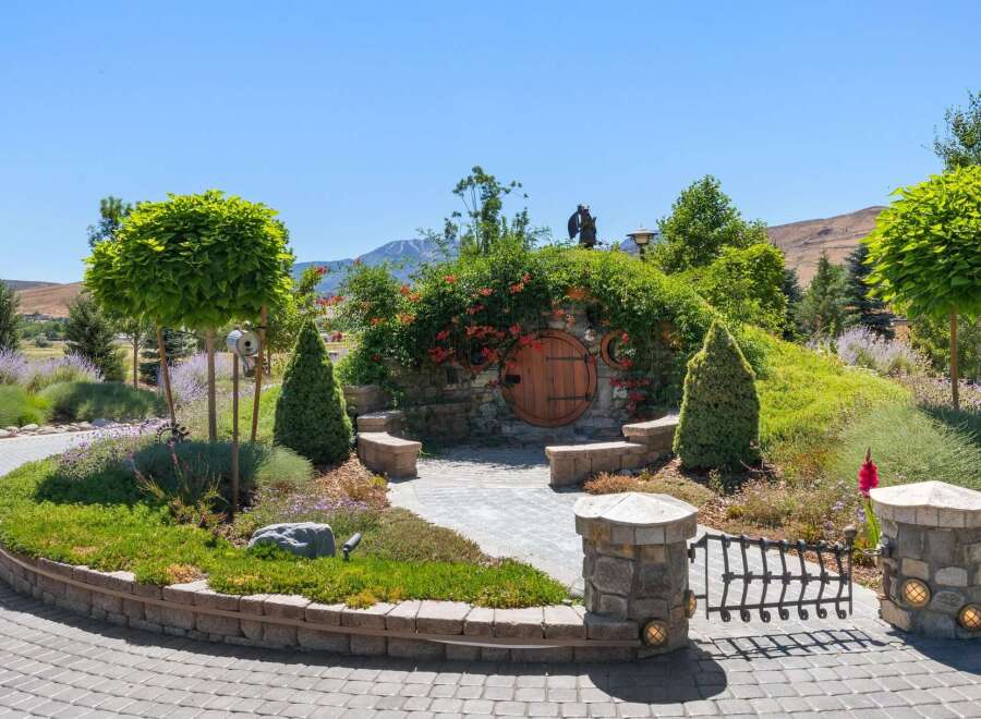 Discover a unique hobbit-inspired home nestled into a hillside, featuring a charming round wooden door and lush landscaping.