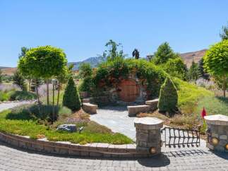 Discover a unique hobbit-inspired home nestled into a hillside, featuring a charming round wooden door and lush landscaping.
