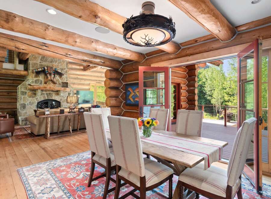Experience rustic elegance in this charming log cabin dining room, featuring a spacious wooden table set for six and a cozy seating area with a stone fireplace.