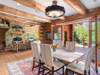 Experience rustic elegance in this charming log cabin dining room, featuring a spacious wooden table set for six and a cozy seating area with a stone fireplace.