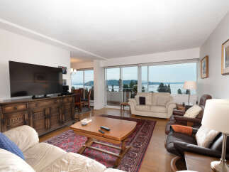 Experience breathtaking water and mountain views from this stunning living room, featuring a large flat-screen TV and a beautiful wooden media console.