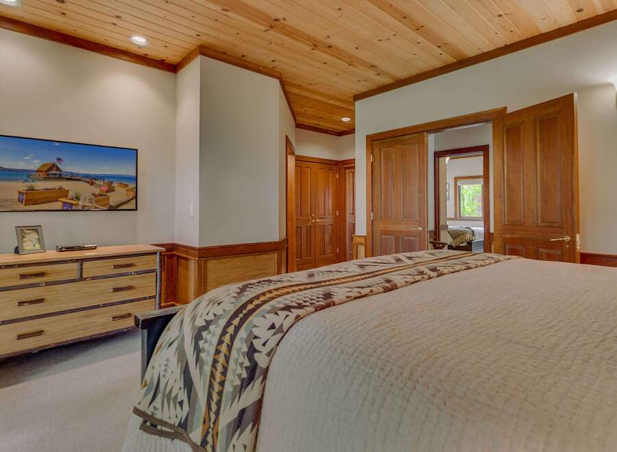 Experience rustic charm in this inviting bedroom, featuring a warm wood-paneled ceiling and a large flat-screen TV mounted above a rustic dresser.