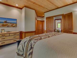 Experience rustic charm in this inviting bedroom, featuring a warm wood-paneled ceiling and a large flat-screen TV mounted above a rustic dresser.