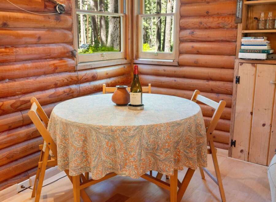 Cozy log cabin dining area featuring a round table draped with a paisley tablecloth, set against a backdrop of warm wood paneling and forest views through the windows.