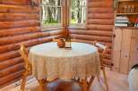 Cozy log cabin dining area featuring a round table draped with a paisley tablecloth, set against a backdrop of warm wood paneling and forest views through the windows.
