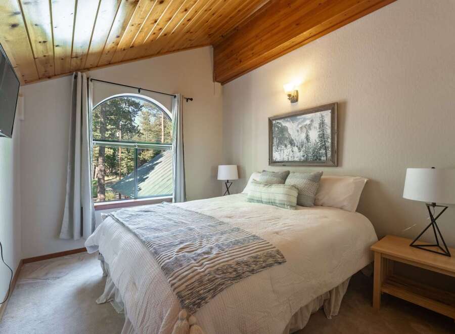 Experience cozy mountain living in this charming bedroom featuring a vaulted wood-paneled ceiling and a large arched window offering picturesque forest views.