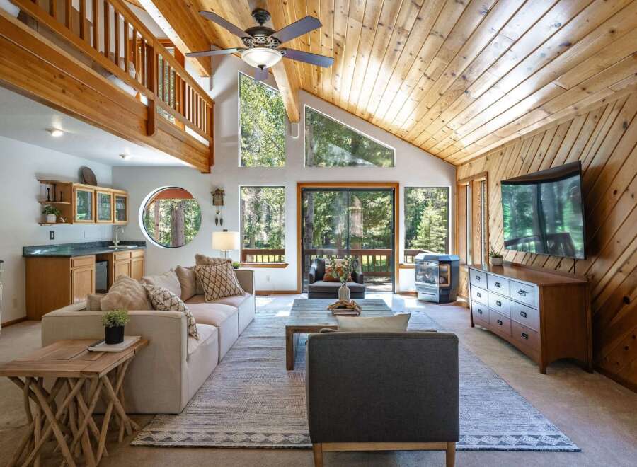 Experience rustic charm in this spacious living room featuring a soaring wood-paneled vaulted ceiling and a wall of windows offering serene forest views.
