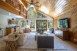 Experience rustic charm in this spacious living room featuring a soaring wood-paneled vaulted ceiling and a wall of windows offering serene forest views.