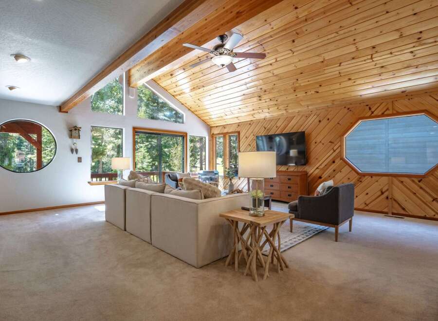 Experience rustic charm and modern comfort in this inviting living room, featuring a vaulted wood-paneled ceiling with a ceiling fan and expansive windows that flood the space with natural light.