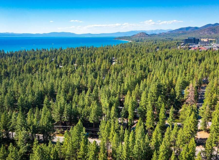 Experience the ultimate lakeside living with this stunning aerial view of a property nestled amidst a lush pine forest, offering breathtaking vistas of a crystal-clear blue lake and distant mountains.
